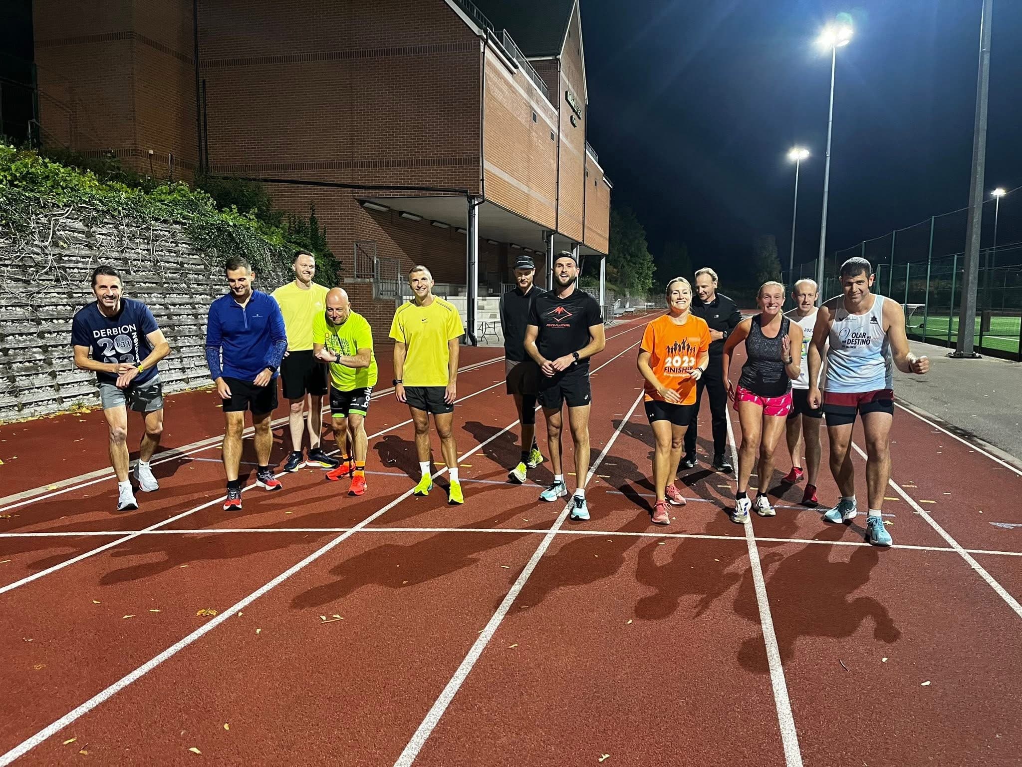 A group of adult club runners standing on a floodlit running track after a session, in the dark